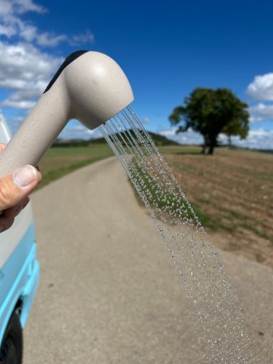 Halter mit sprudelndem Wasser, im Hintergrund ein Baum auf einem Feldweg.
